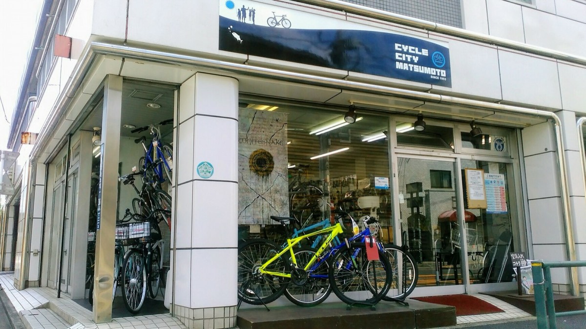 紺色の看板に「CYCLE CITY MATSUMOTO」と書かれた、角にある白い外壁の店舗。店先にクロスバイクが並んでいる